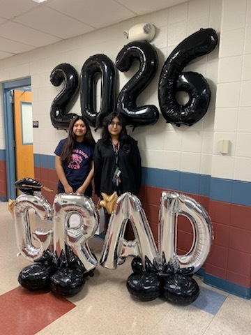 Two students stand smiling in front of a school hallway. Behind them are large, shiny balloons spelling "2026 GRAD" in black and silver. The mood is celebratory.
