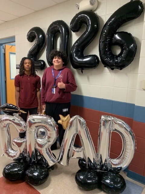 Two students stand in front of large black "2026" balloons and silver "GRAD" balloons. One gives a thumbs up, conveying a celebratory tone.