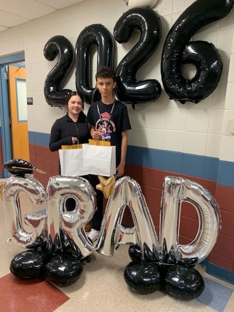 Two students stand in a hallway with large "2026" and "GRAD" balloon displays. They hold gift bags, conveying a celebratory graduation tone.