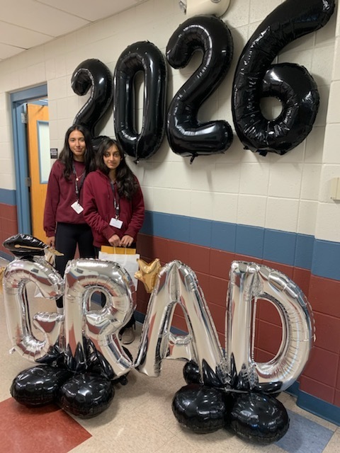 Two people in maroon sweaters stand smiling next to large "2026" balloons and silver "GRAD" balloons in a school hallway.