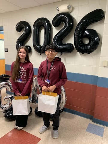 Two students stand smiling in a school hallway, holding gift bags. Behind them, large black balloons spell out "2026," celebrating their graduating class.
