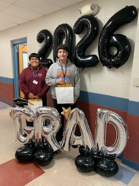 Two students smiling in a hallway stand between large black balloons forming "2026" and silver balloons spelling "GRAD," holding gift bags.