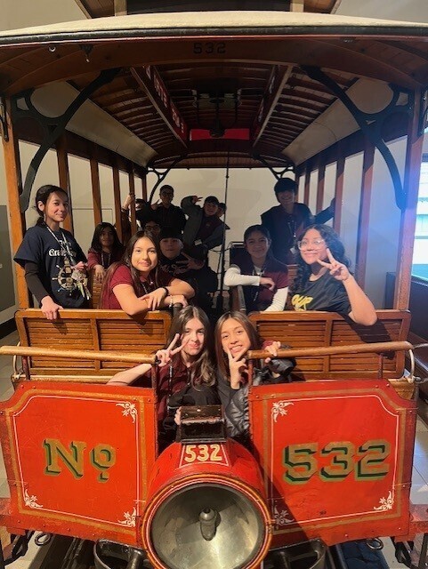A group of smiling children pose inside a vintage red trolley marked "No. 532," flashing peace signs and expressing joy and excitement.