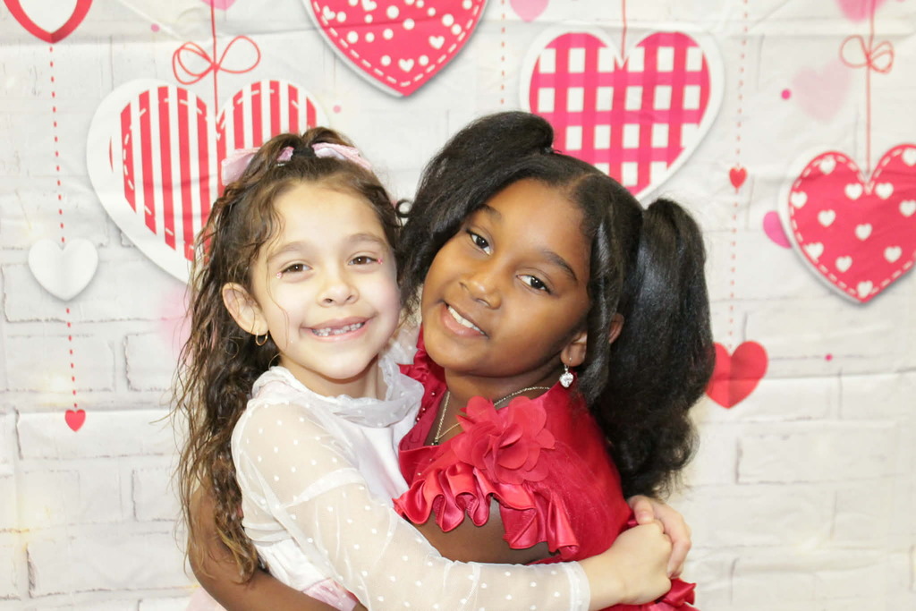 Two smiling girls in pink and red dresses hug in front of a Valentine's-themed backdrop with hanging heart decorations, conveying joy and friendship.