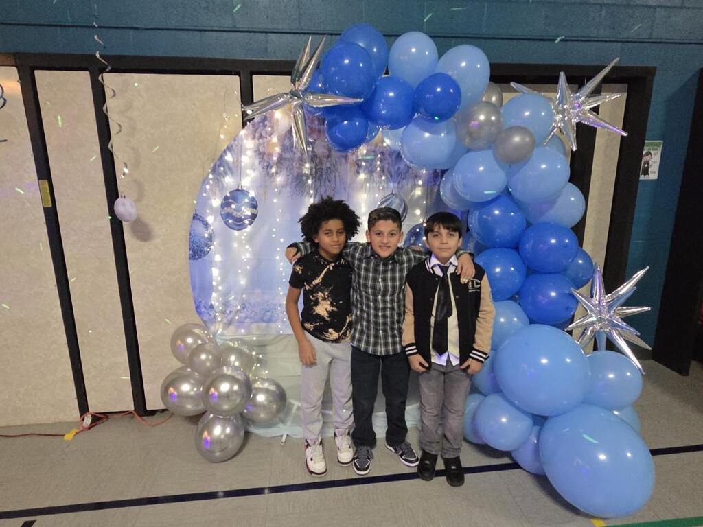 Three children stand together smiling in front of a festive backdrop with blue and silver balloons and star decorations, creating a joyful atmosphere.