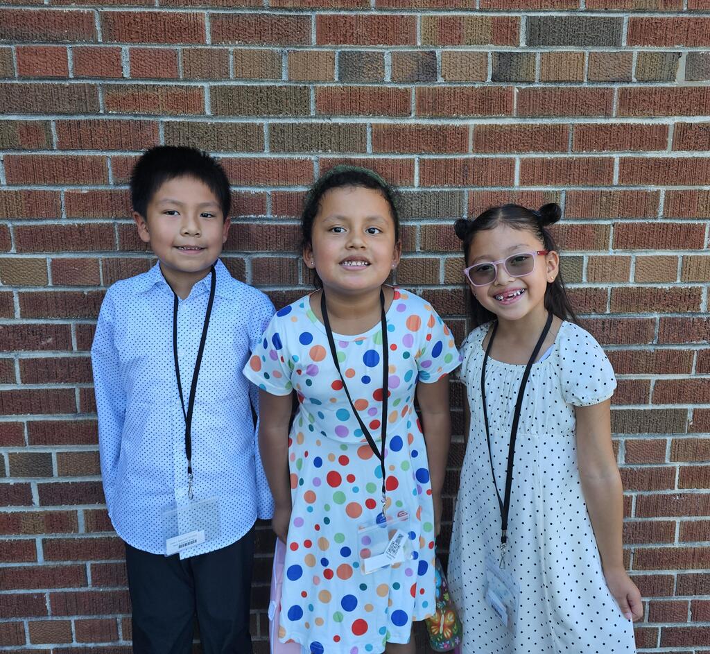 Three children stand smiling in front of a brick wall, wearing lanyards. The left child wears a blue shirt, the center a polka-dot dress, and the right a white dress.