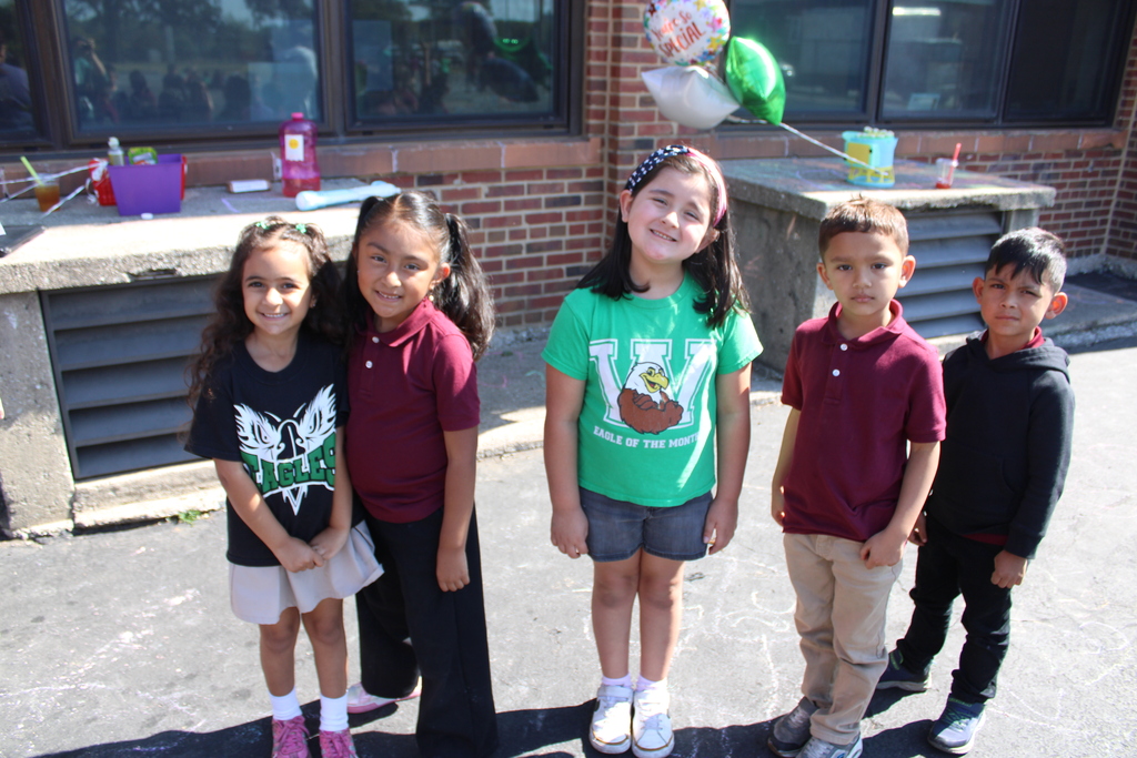Five children smiling outside in front of a brick wall with windows. They wear casual clothes; balloons and chalk drawings are in the background.