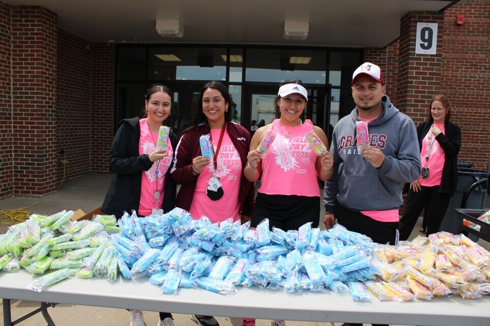 A group of four individuals, dressed in similar pink shirts, are smiling while holding ice pops. They are standing behind a table piled with colorful ice pops in various shades.