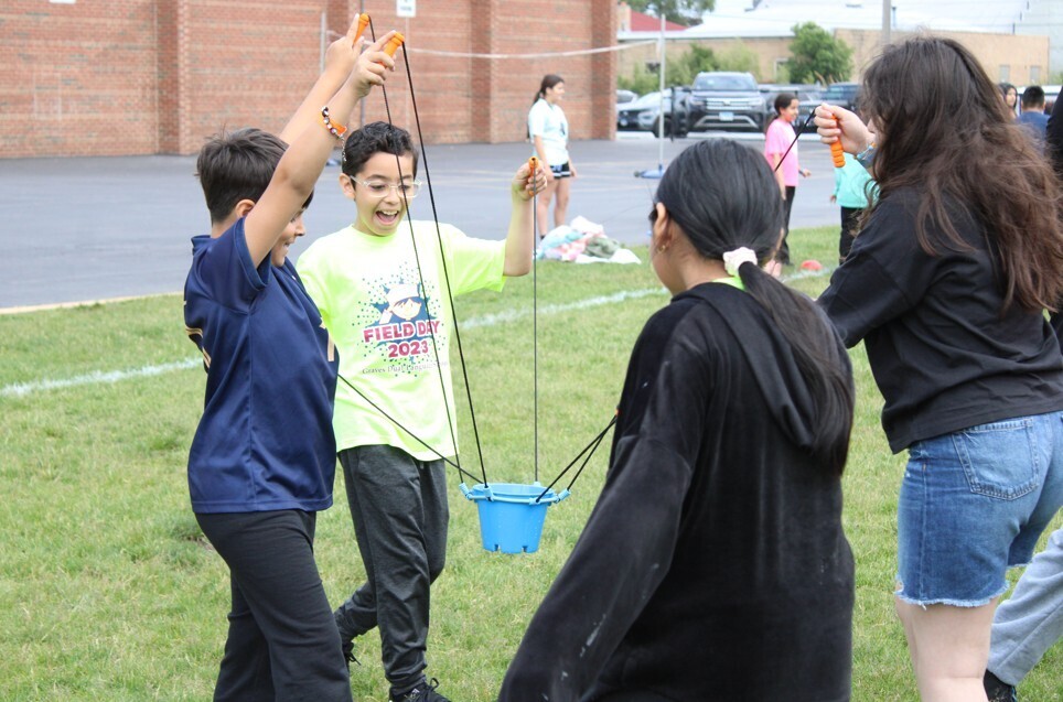 Children participating in a field day activity, working together to lift a bucket using ropes.