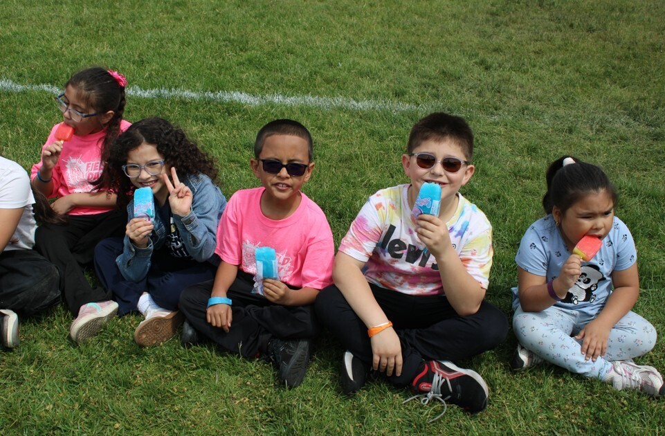 A group of children sitting on grass, holding popsicles. They are smiling and wearing casual clothing, with some kids wearing sunglasses.