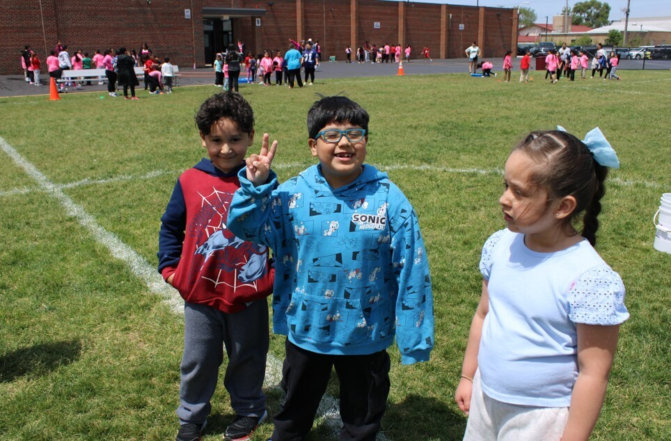 Three children are standing on a grassy field. The child in the middle is wearing glasses and a blue hoodie with a graphic design, making a peace sign. The child on the left is wearing a red Spiderman sweatshirt, and the child on the right is in a light blue shirt with her hair tied back. In the background, there are groups of children playing and participating in activities.