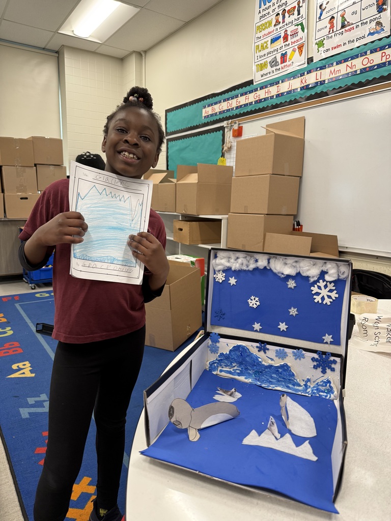 A smiling young girl holds a drawing next to a diorama depicting an arctic scene with a seal and icebergs.