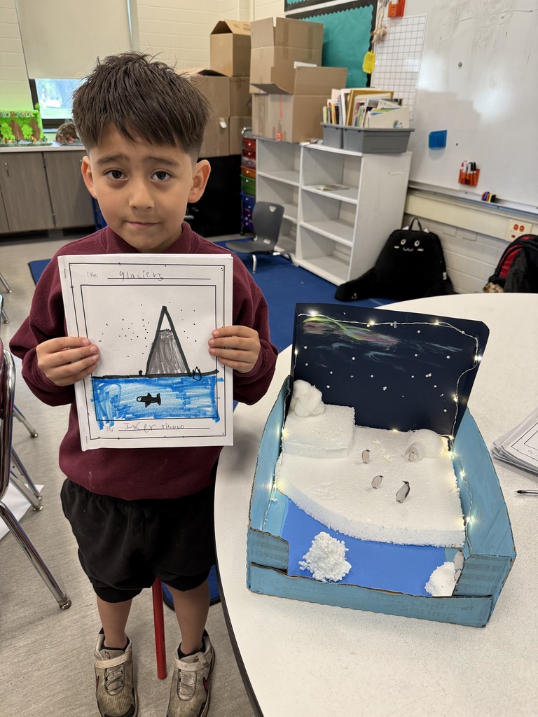 Young student proudly displays a drawing of a glacier next to a three-dimensional diorama depicting an icy arctic scene with penguins and string lights.