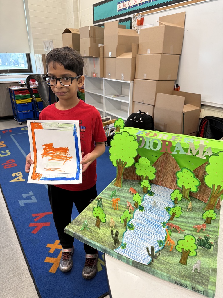Young boy in glasses stands proudly holding a drawing next to his three-dimensional forest diorama featuring miniature animals and a drawn river.