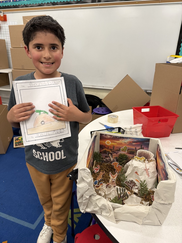 Smiling young student holding a drawing stands next to his shoebox diorama depicting desert animals and landscape.