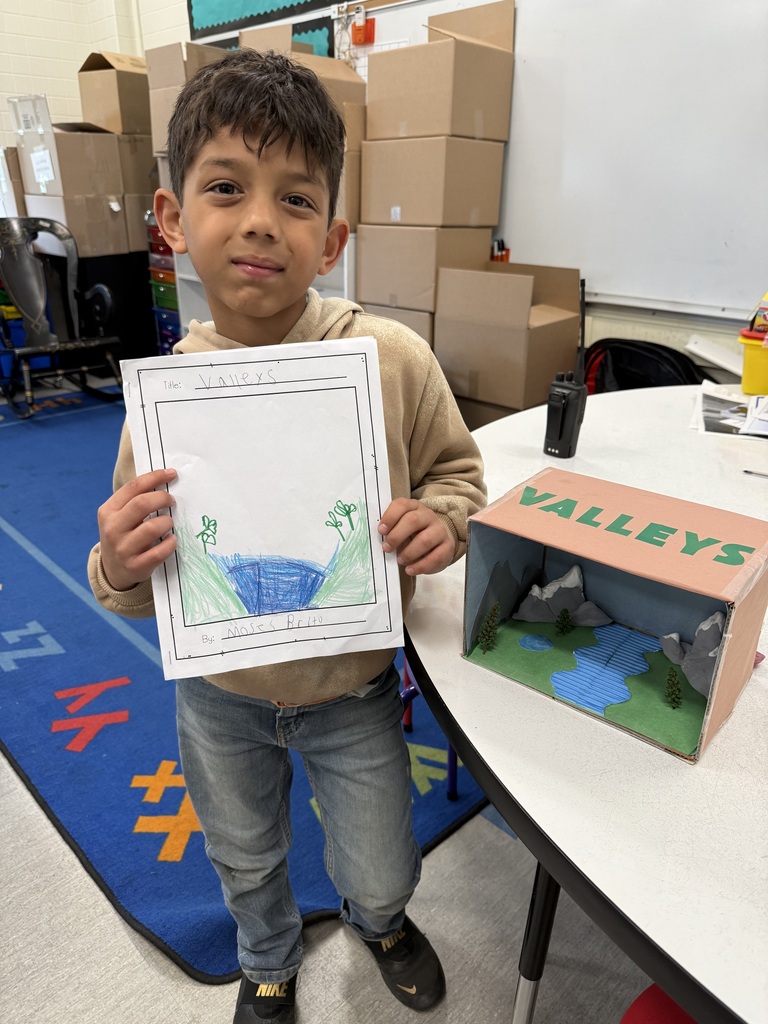 Young student proudly displays his drawing and diorama project illustrating a valley in a classroom setting.