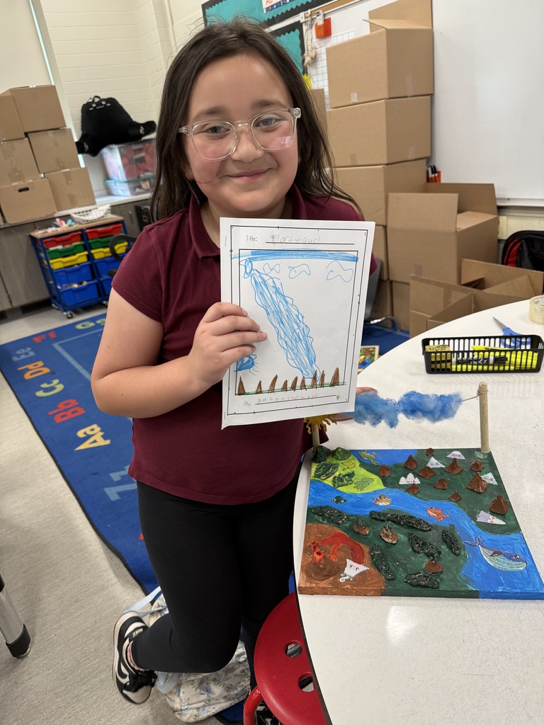 Smiling elementary student in glasses proudly displays her water cycle drawing next to a three-dimensional landscape model in a classroom setting.