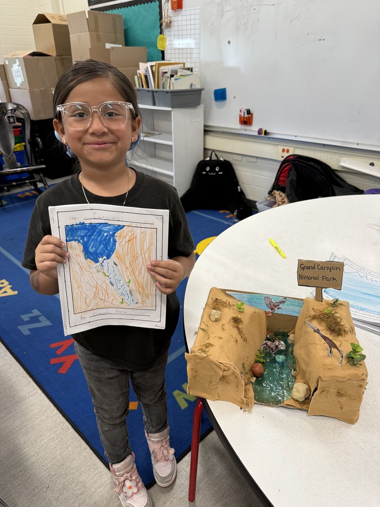 Smiling girl in glasses holds a drawing next to her 3D Grand Canyon National Park diorama project in a classroom setting.