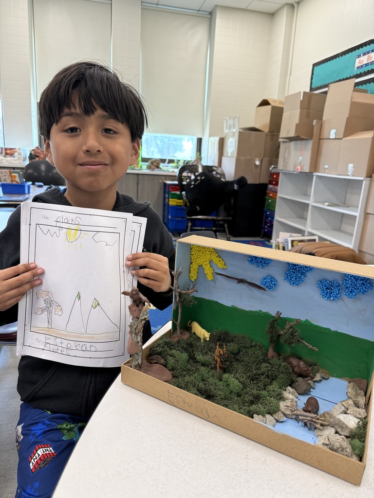 A smiling young boy stands in a classroom holding a drawing labeled "Plains" next to a diorama featuring mossy terrain, small animal figures, and a river.