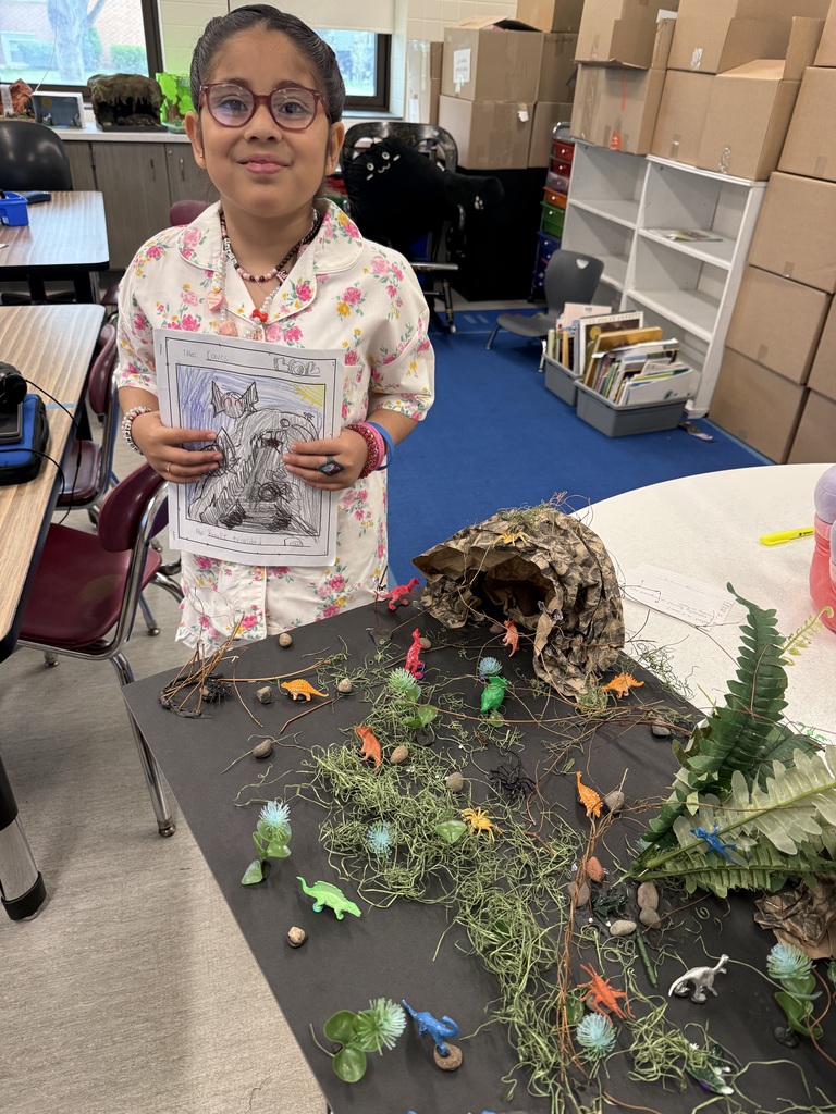 Young girl in pajamas displays a drawing next to a diorama featuring small plastic dinosaurs, moss, and a cave structure in a classroom setting.