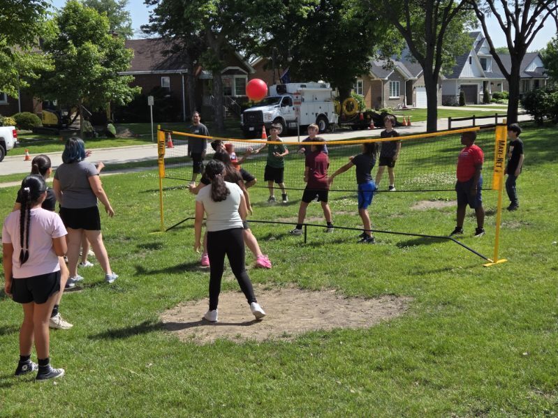 A group of children and adults play outdoor volleyball with a red ball over a low net on a sunny grassy area in a residential neighborhood.