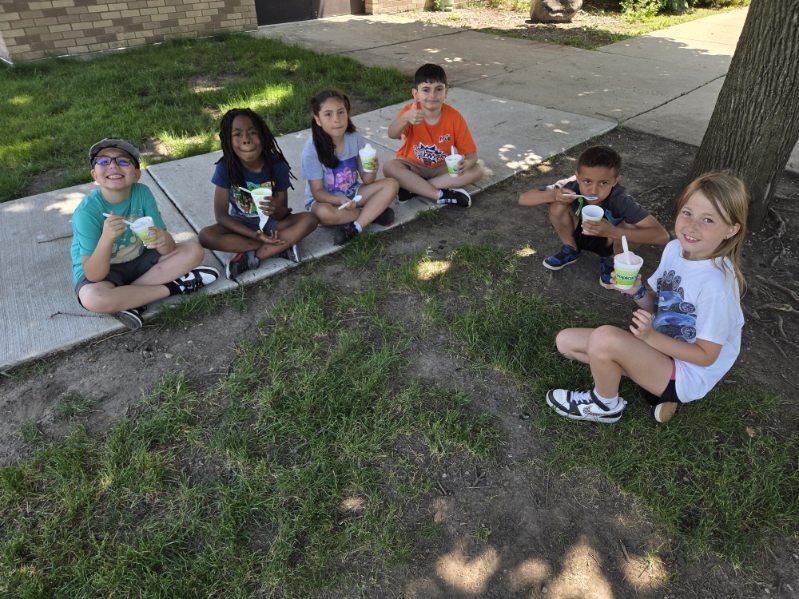 Six diverse children sit outdoors on a sunny day, enjoying ice cream or frozen treats near a concrete sidewalk and grassy area.