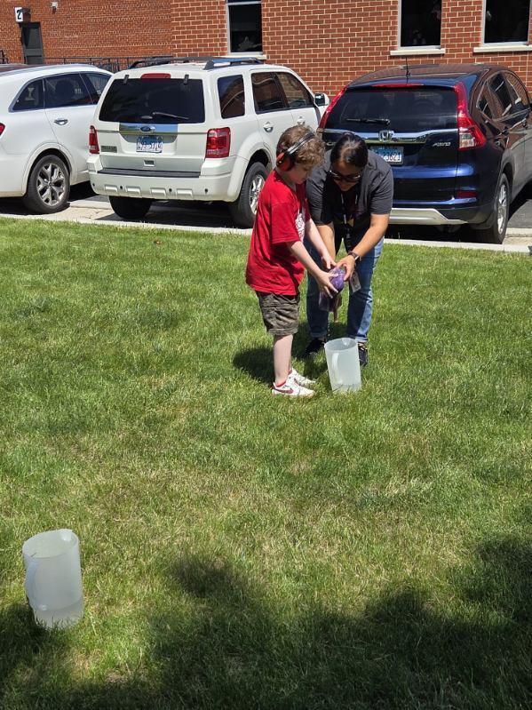 A young boy wearing headphones and a red shirt works with an adult outdoors on a sunny day to wring a purple cloth over a plastic pitcher on a grassy lawn near parked cars and a brick building.