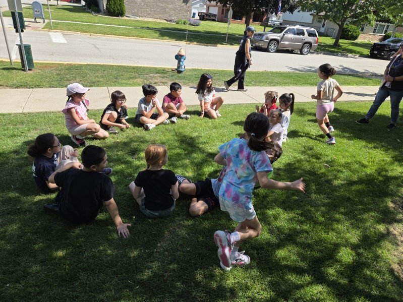 A group of diverse young children sits and stands on a sunny green lawn near a suburban street while an adult supervises.