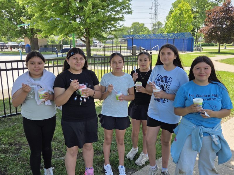 Six young girls stand outdoors in a park setting, enjoying cups of shaved ice or ice cream on a sunny day.