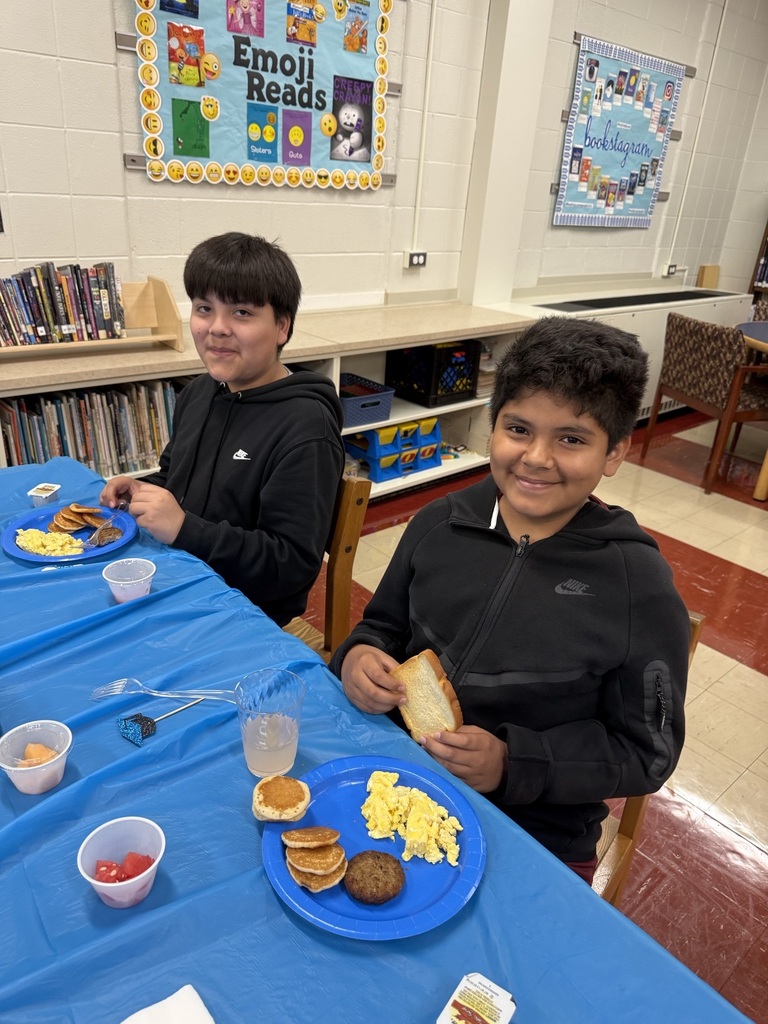 Four diverse elementary school-aged girls sit at a table covered with a bright blue tablecloth enjoying breakfast in a classroom setting.