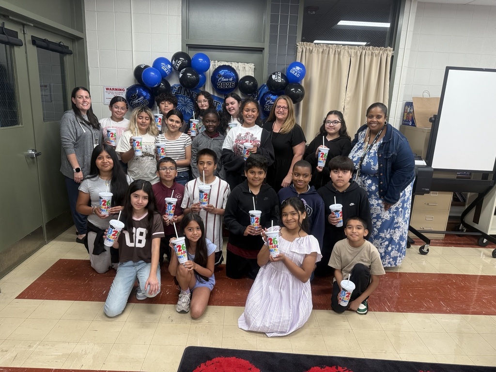 A group of diverse students and female teachers pose together indoors holding matching white and red branded cups, with blue and black graduation balloons visible in the background.