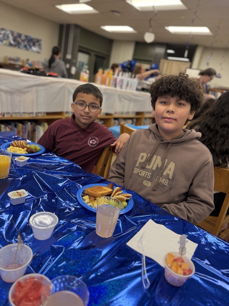 Two boys sit at a table covered with a blue shiny cloth, eating breakfast in what appears to be a school library setting.