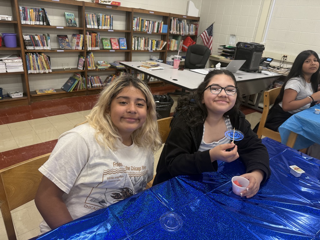 Four diverse elementary school-aged girls sit at a table covered with a bright blue tablecloth enjoying breakfast in a classroom setting.