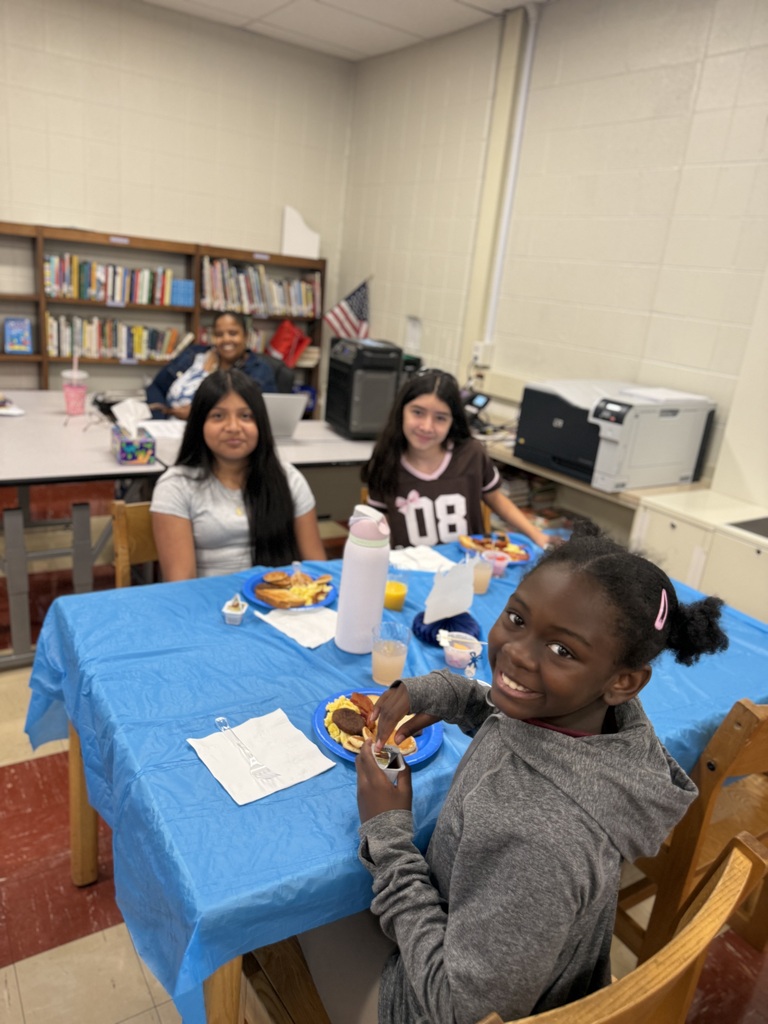 Four diverse elementary school-aged girls sit at a table covered with a bright blue tablecloth enjoying breakfast in a classroom setting.