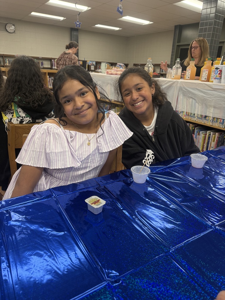 Two smiling young girls sit at a table covered with a bright blue, shiny tablecloth inside a room lined with bookshelves.