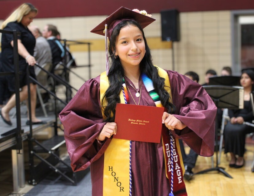 Smiling graduate wearing a maroon cap and gown with honor cords holds a red diploma folder at a school ceremony.