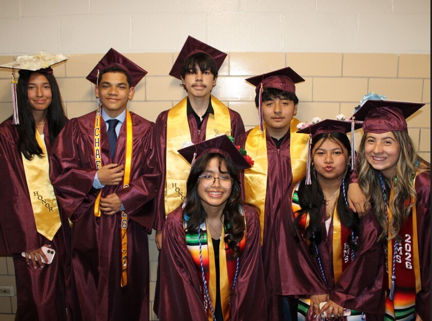 Seven diverse high school graduates wearing maroon gowns, mortarboards, and gold honor stoles pose together indoors against a light-colored cinder block wall.
