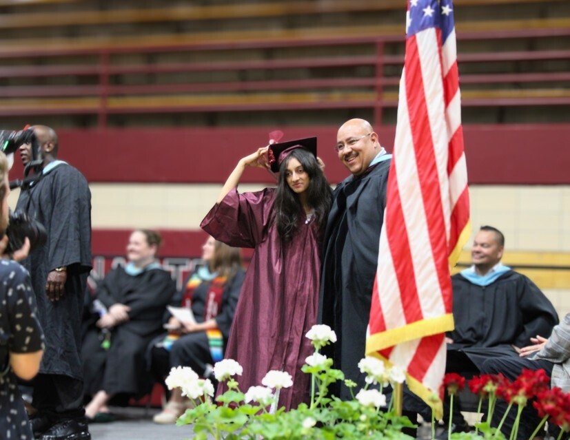 A smiling female graduate in a maroon gown adjusts her cap beside a male administrator next to an American flag during a graduation ceremony indoors.