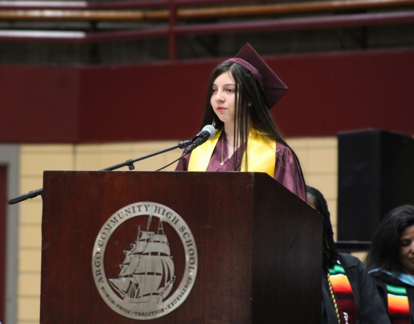A female high school student wearing a maroon cap and gown with a yellow stole speaks at a podium bearing the Argo Community High School logo during a graduation ceremony.