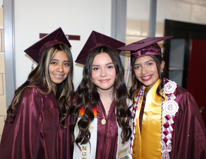 Three smiling female graduates wearing maroon gowns and caps pose together indoors, with one student wearing a gold stole and braided cords.
