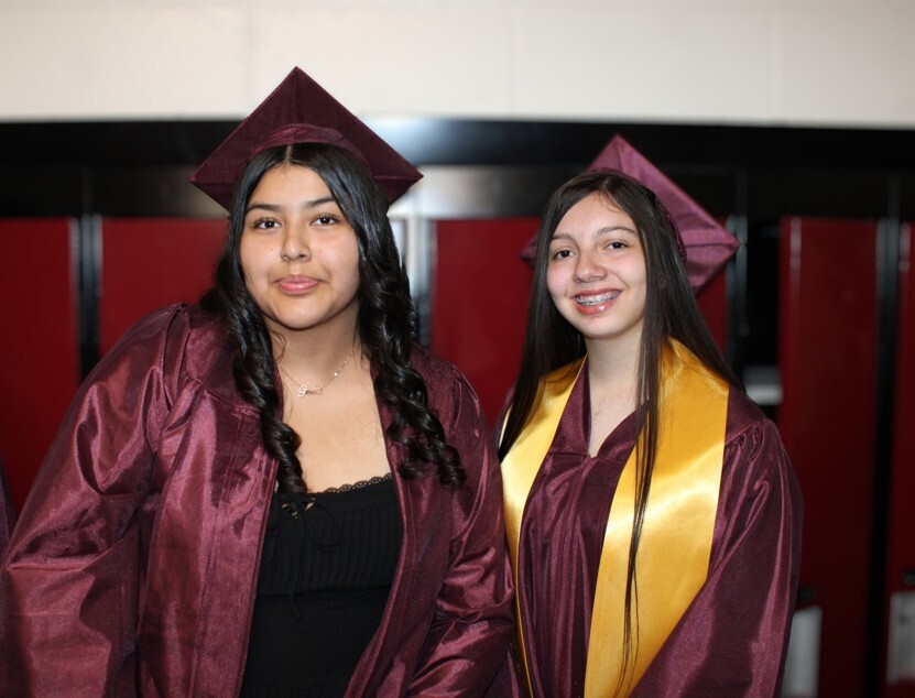 Two young women in maroon graduation gowns and caps stand side-by-side, one wearing a gold stole, against a background of dark red panels.