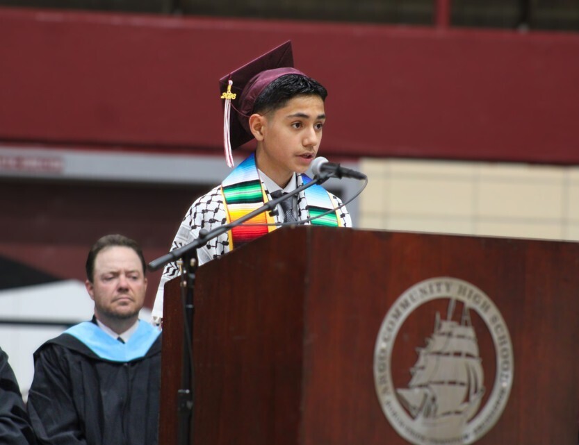 A male high school graduate wearing a graduation cap and a Keffiyeh and rainbow sash speaks at a wooden podium featuring a argo Community High School logo while an administrator in academic regalia looks on.