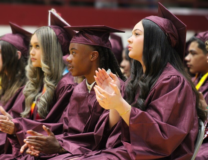 Two young women in maroon graduation gowns and caps stand side-by-side, one wearing a gold stole, against a background of dark red panels.