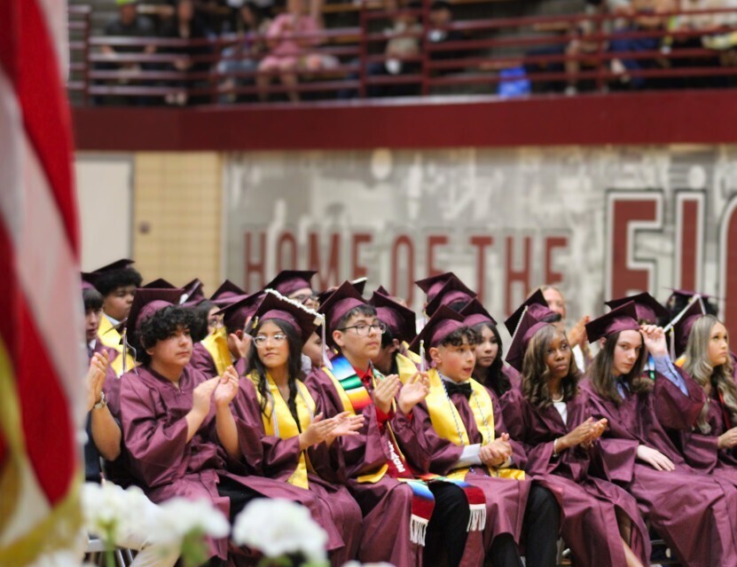 Row of diverse high school graduates in maroon caps and gowns applaud during an indoor commencement ceremony beside a blurred American flag.