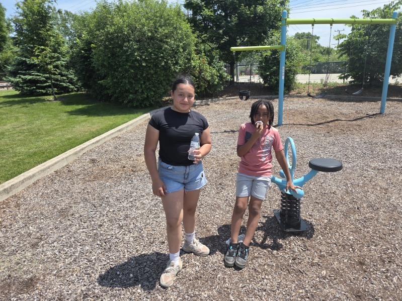 Two grade school kids stand in a playground with a swing set and a spring rider.