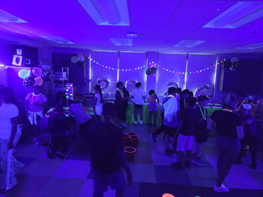 Children wearing neon glow sticks and necklaces dance in a dimly lit room illuminated by blacklights at a party.