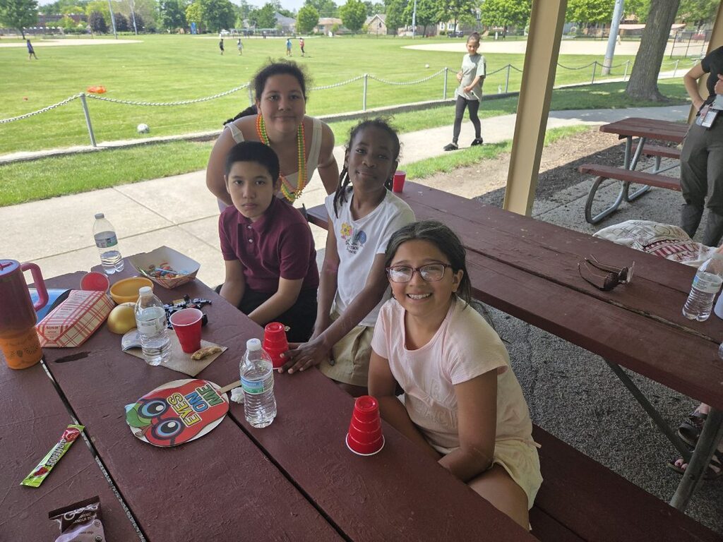 Four children sit at a picnic table outdoors with food and drinks, a grassy field with other children playing in the background.