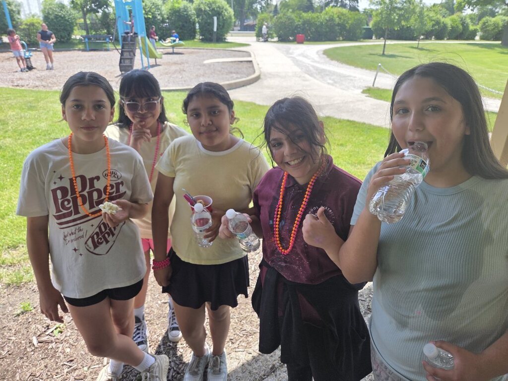 Five young girls wearing colorful necklaces and casual outfits stand together outdoors near a playground.
