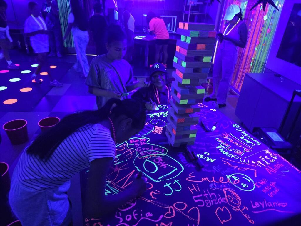 Children and adults in a blacklight room with neon drawings on a table and a tower of Jenga blocks.