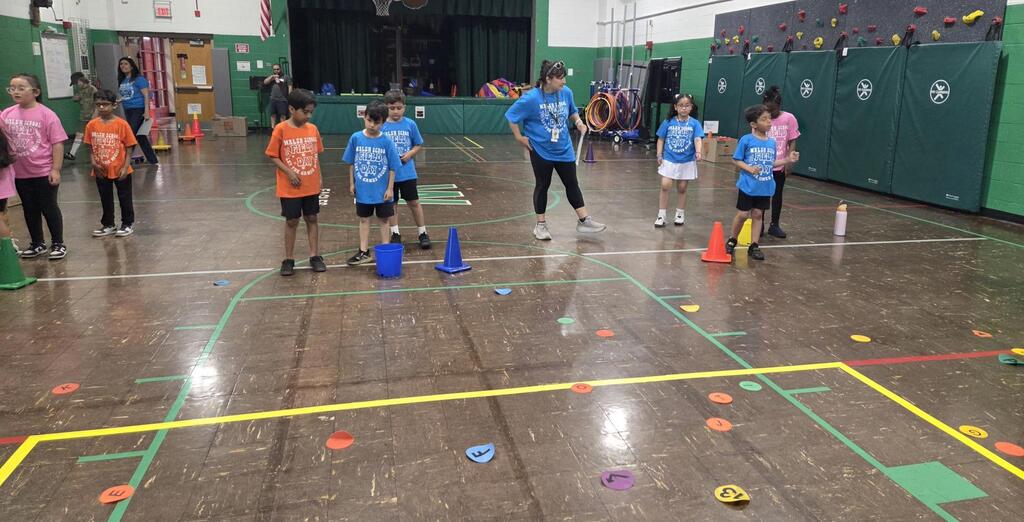 Children participate in a gym class obstacle course with colorful markers, cones, and adults overseeing the activity.